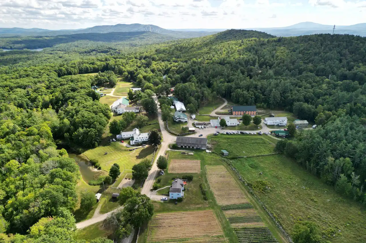 The His Mansion campus from the air, white buildings along a dirt road above planted fields, with wooded hills behind