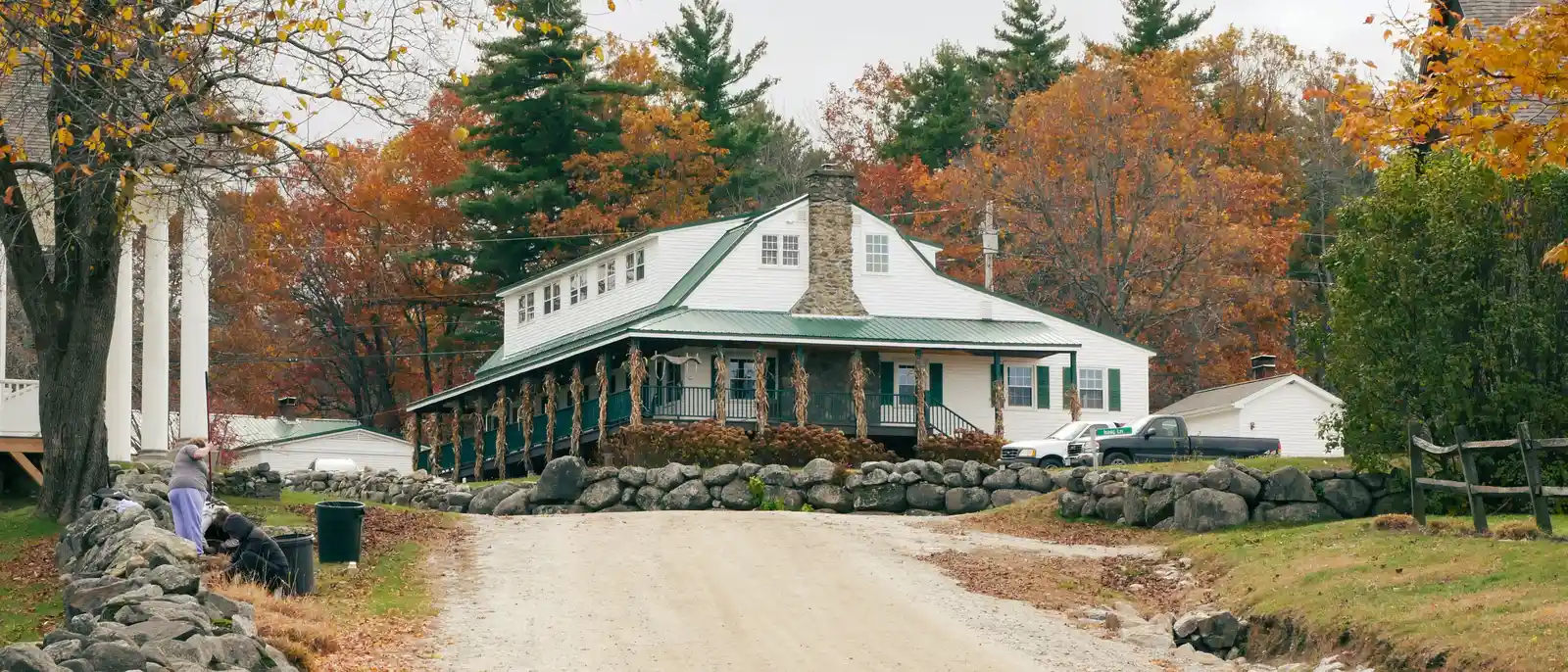White building with a stone chimney and green wraparound porch at the end of a gravel road, framed by autumn maples