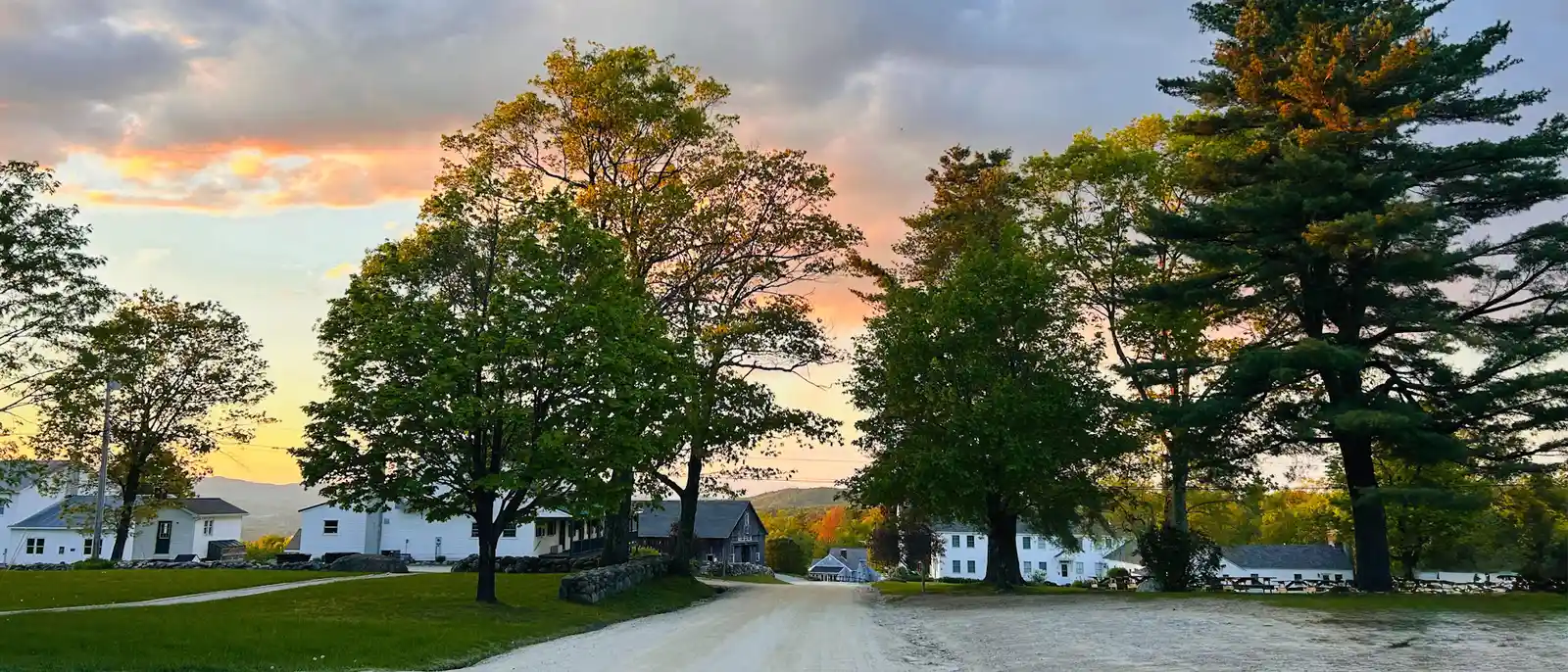 Dirt road through the His Mansion campus at sunset, with tall maples and pines beside white clapboard buildings