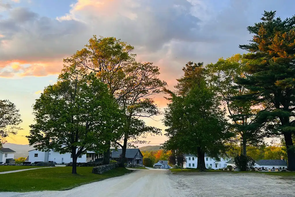 Dirt road through the His Mansion campus at sunset, with tall maples and pines beside white clapboard buildings