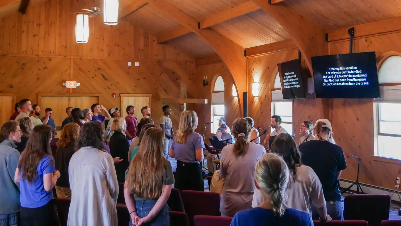 A full chapel with timber beams stands for worship, facing a band and two screens showing song lyrics