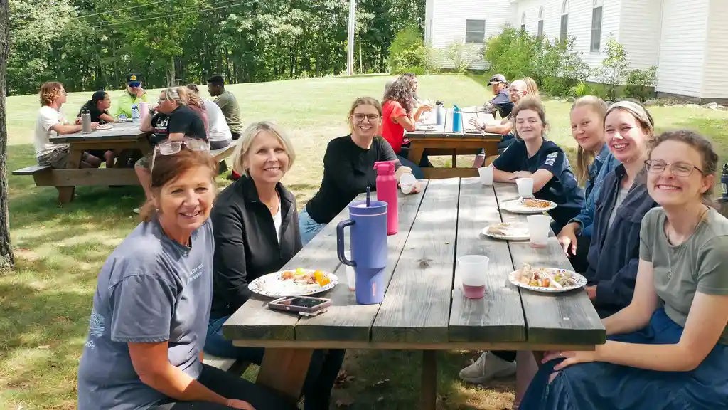 Women smiling at a picnic table with paper plates and cups, with more people eating at tables behind them