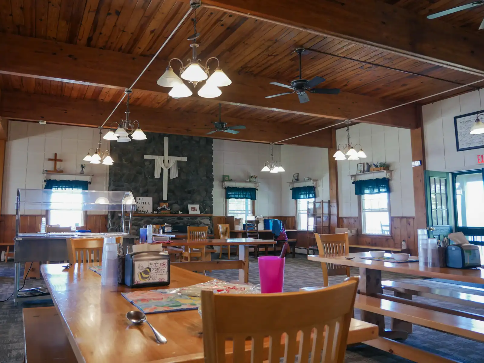 Long wooden tables and chairs in Griggs Dining Hall under a pine ceiling and chandeliers, with a white cross on a stone wall