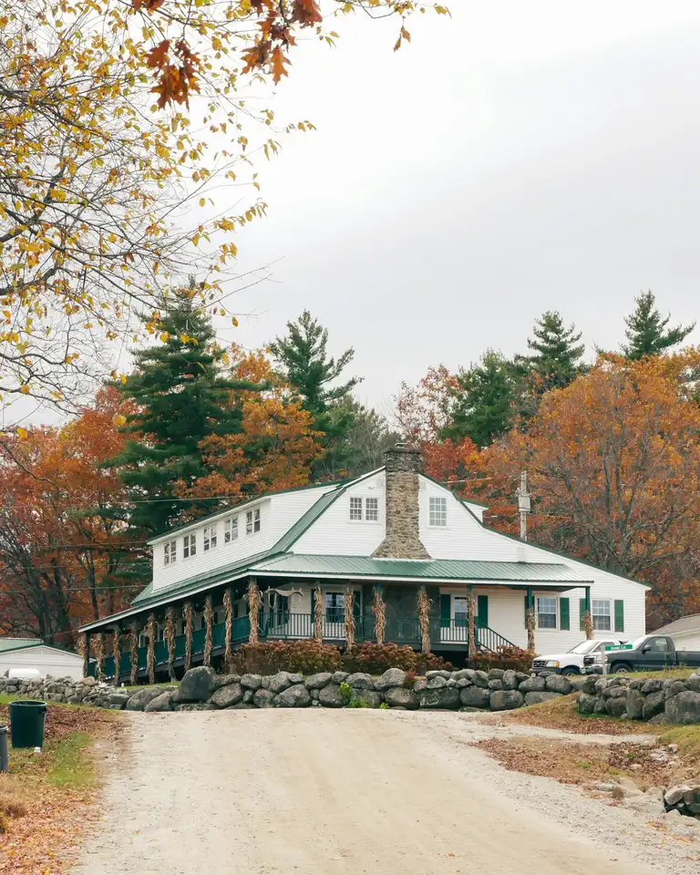 The same white house in fall, orange and red trees behind it and yellow leaves overhead, under a gray sky