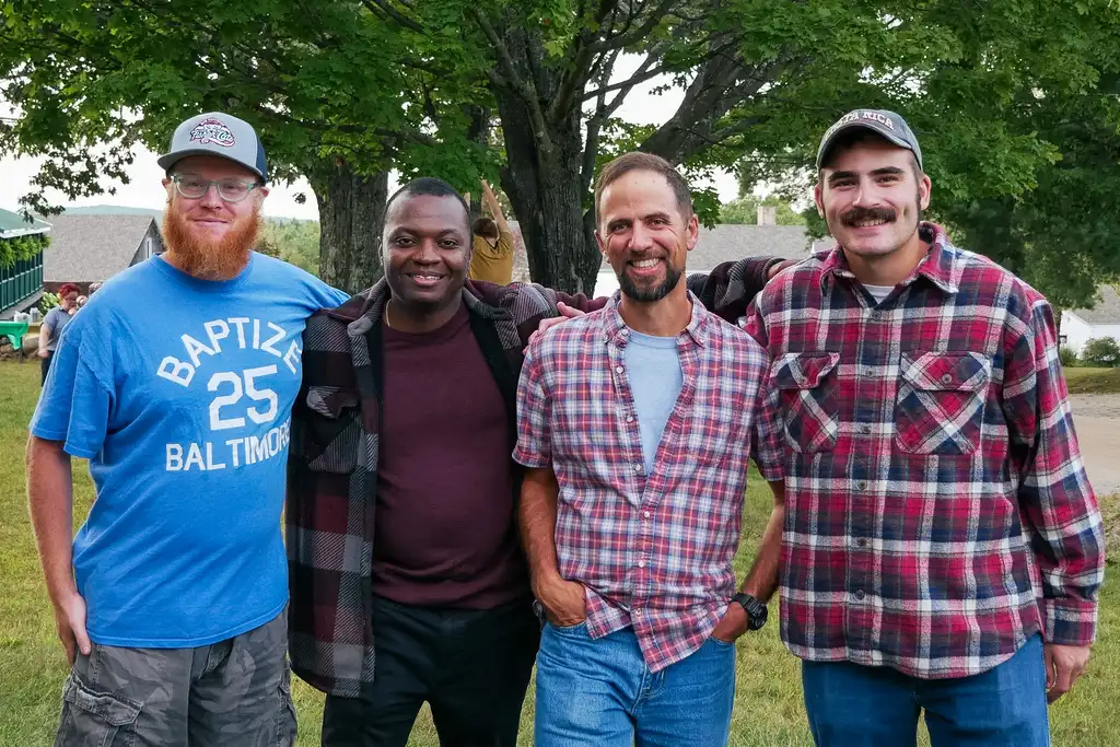 Four smiling men stand arm in arm on a lawn under a large tree, three of them in plaid shirts