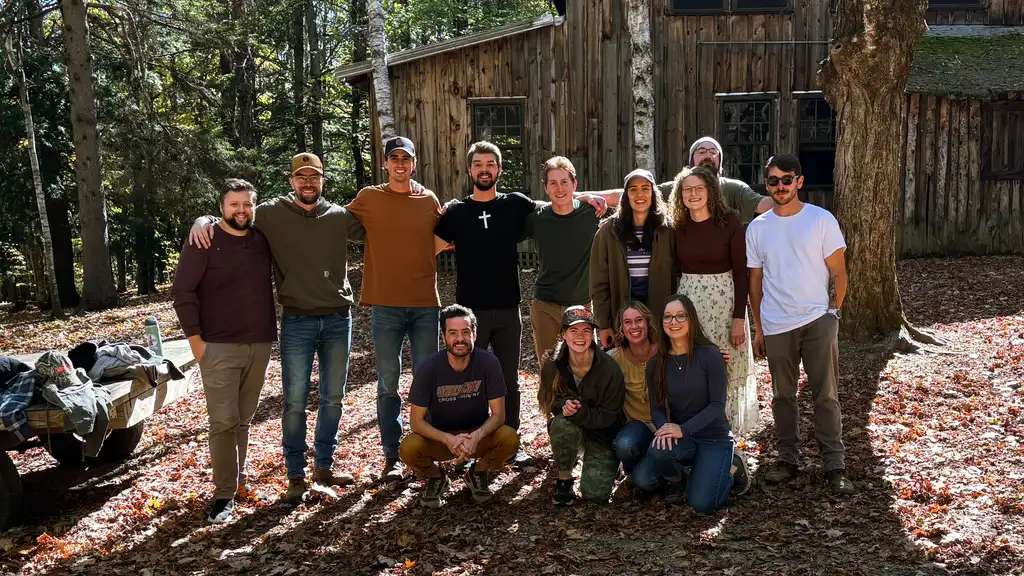 Thirteen smiling young adults, some kneeling, in front of a weathered wooden building in a sunlit autumn forest