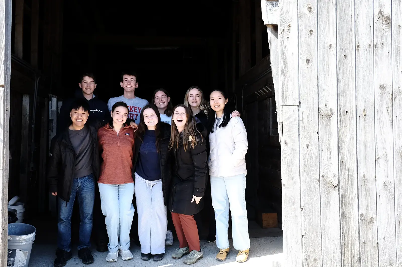 Ten smiling young volunteers standing together in the open doorway of a weathered wooden barn